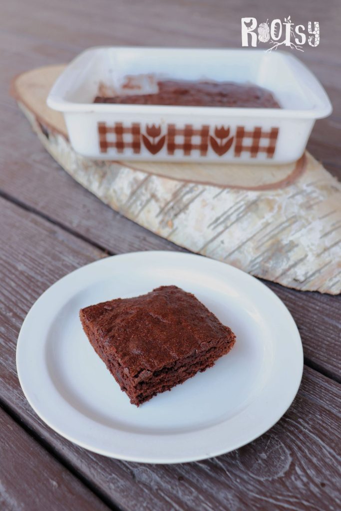 A brownie sits on a plate in front of a board with remaining pan of brownies on top behind it.