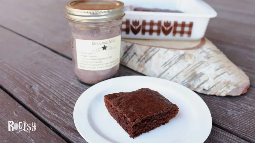 A brownie sits on a plate. In the background is a jar of brownie mix and the remaining brownies in the pan.