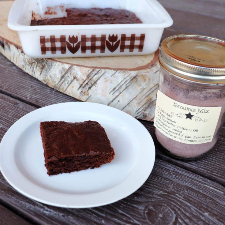 A brownie on a plate with pan of brownies in the background. To the left a jar with brownie mix inside.