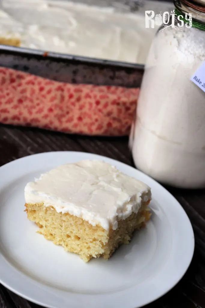 A slice of white cake with white frosting sits on a plate in front of a jar and red printed cloth.