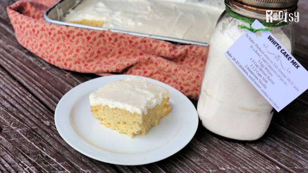 A slice of white cake with white frosting sits on a plate. To the right is a canning jar labeled 'white cake mix'. In the background is a metal pan full of cake.