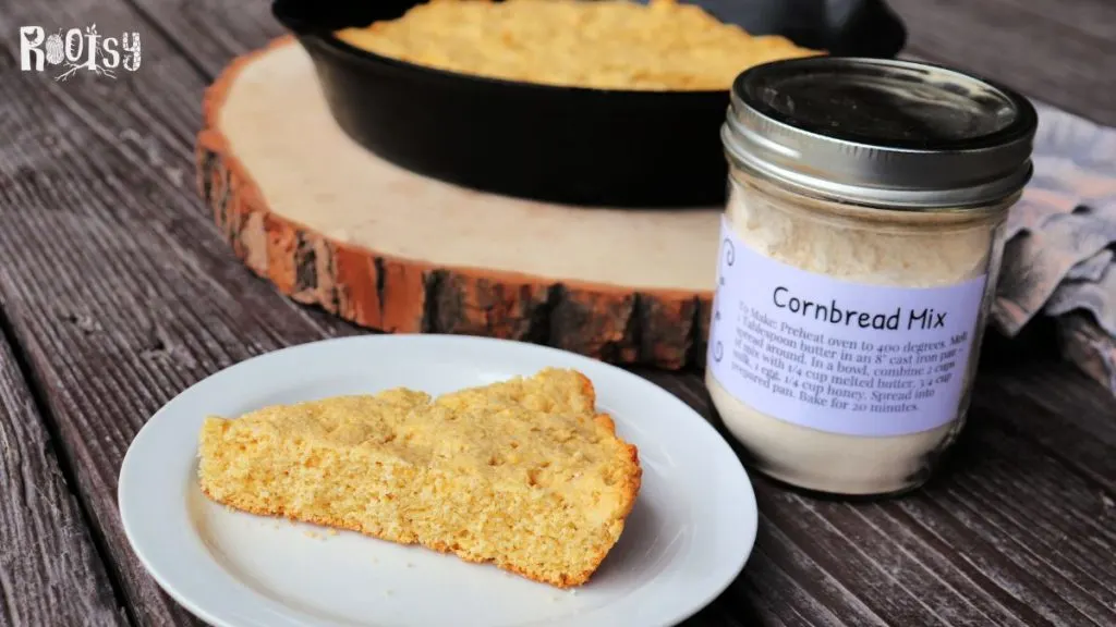 A slice of cornbread sits on a plate in front of a jar full of cornbread mix and a skillet of baked cornbread.