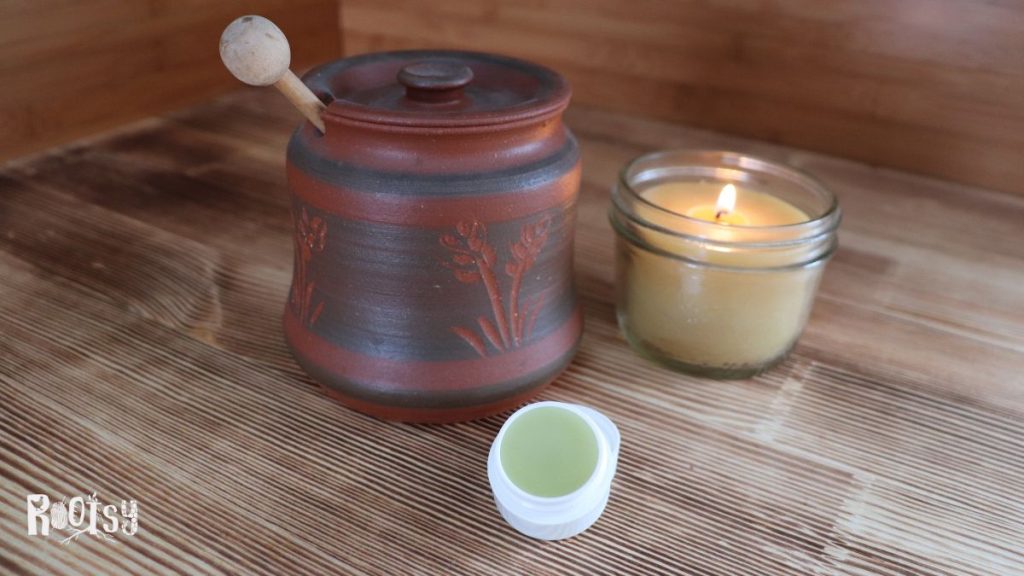 A container of lip balm sits in front of a clay honey jar and lit beeswax candle.