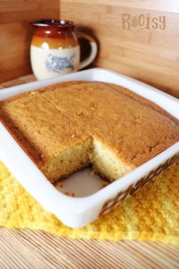 A square baking dish with a piece of cornbread removed, showing its golden-brown crust. The dish sits on a yellow textured cloth, and a ceramic mug with a brown rim is in the background.