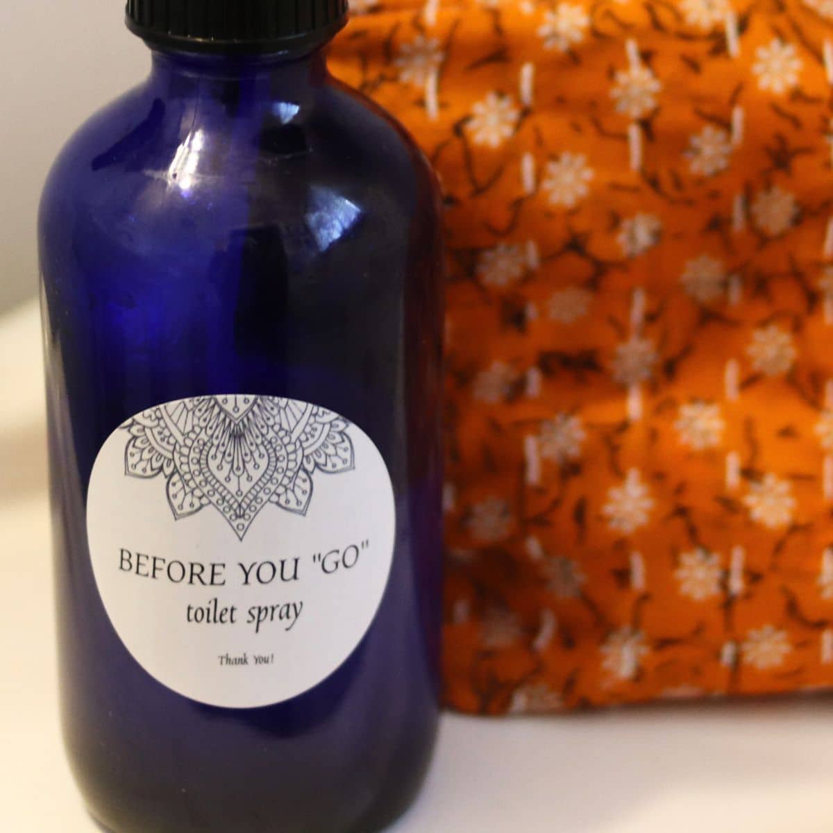 Blue bottle labeled "Before You 'Go' Toilet Spray" with a decorative pattern, placed next to an orange fabric with a white floral design.