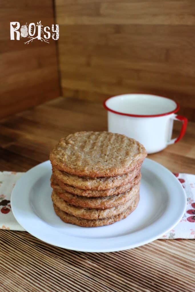A stack of six peanut butter cookies sits on a white plate with a white mug that has a red rim in the background. The setting features a wooden table and a wooden wall, with a napkin under the plate.
