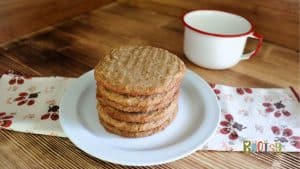 A stack of six round cookies sits on a white plate, placed on a floral-patterned napkin. Behind the plate is a white enamel cup with a red rim, all set on a wooden surface.