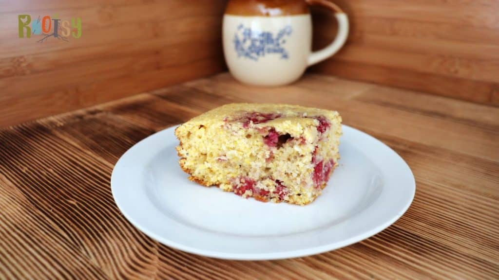 A slice of cornbread with visible pieces of raspberries is served on a white plate. The plate sits on a wooden surface, and there is a ceramic mug with a handle in the background.
