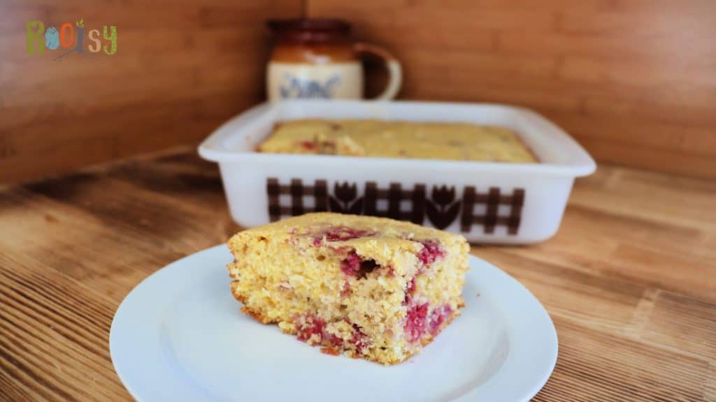 A slice of raspberry cornbread sits on a white plate in the foreground, with the remaining cornbread in a white baking dish behind it. The background features a wooden surface and a ceramic mug.