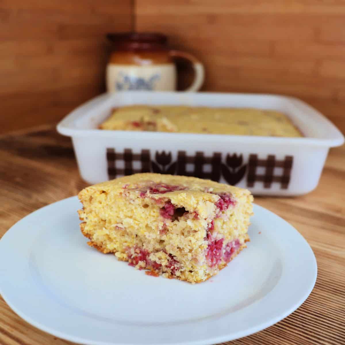 A square piece of raspberry cornbread sits on a white plate in the foreground, with a larger baking dish of cornbread and a brown ceramic mug in the background on a wooden surface.
