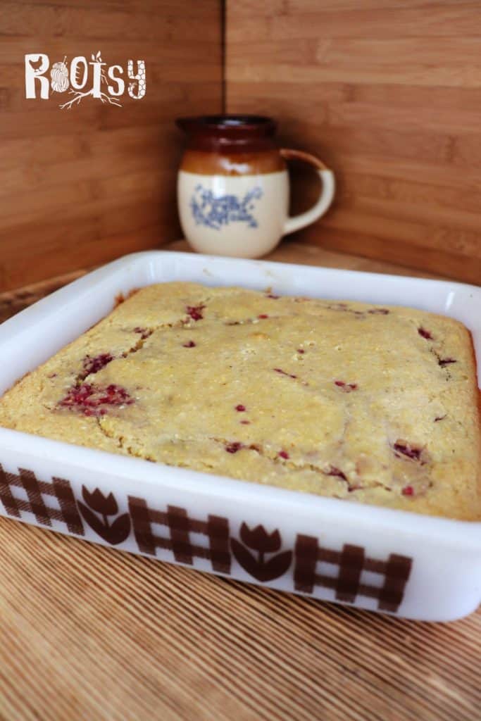 A square baking dish with a golden-brown cornbread featuring raspberry pieces sits on a wooden surface. In the background, there is a ceramic mug with a brown rim and handle. The word "Rootsy" appears at the top.