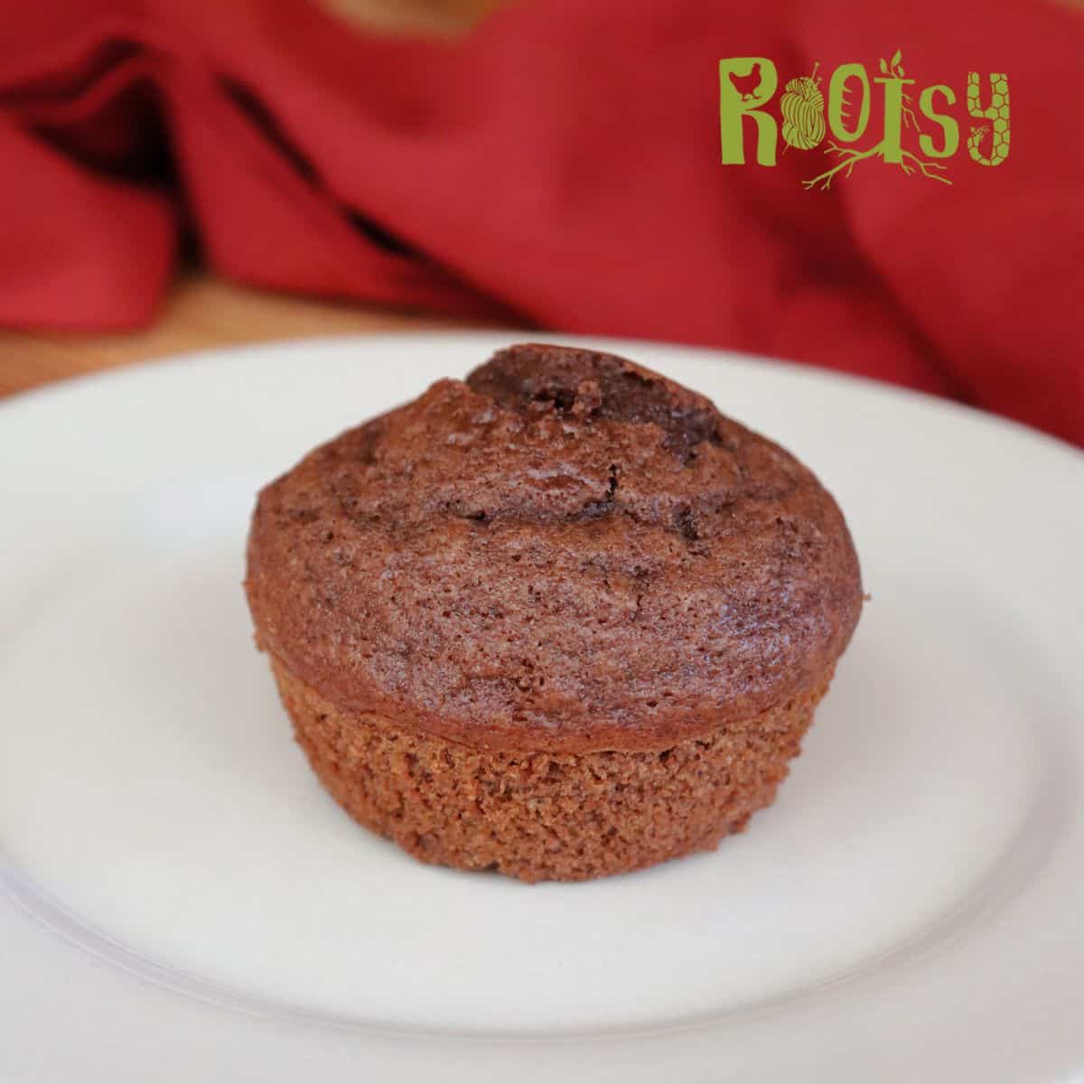 A single chocolate muffin sits on a white plate with a red cloth in the background. The word "Rootsy" appears in green in the top right corner of the image.