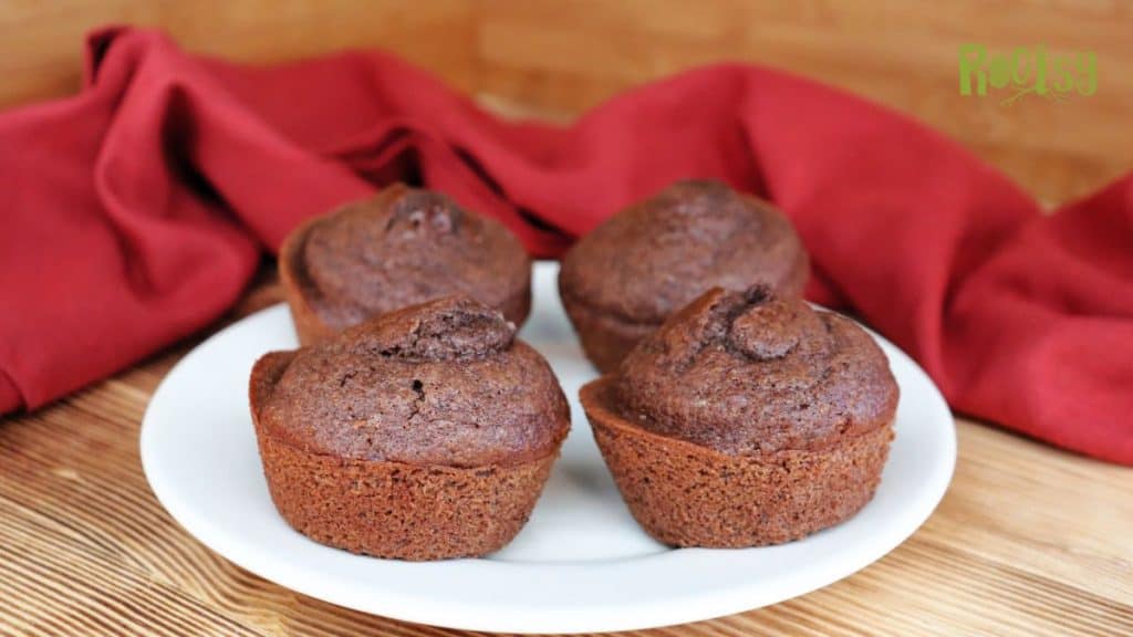 Four chocolate muffins on a white plate, set on a wooden surface with a red cloth in the background.