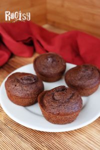 Four chocolate muffins on a white plate, set on a wooden surface with a red cloth in the background and the word "Rootsy" in white text at the top left corner.