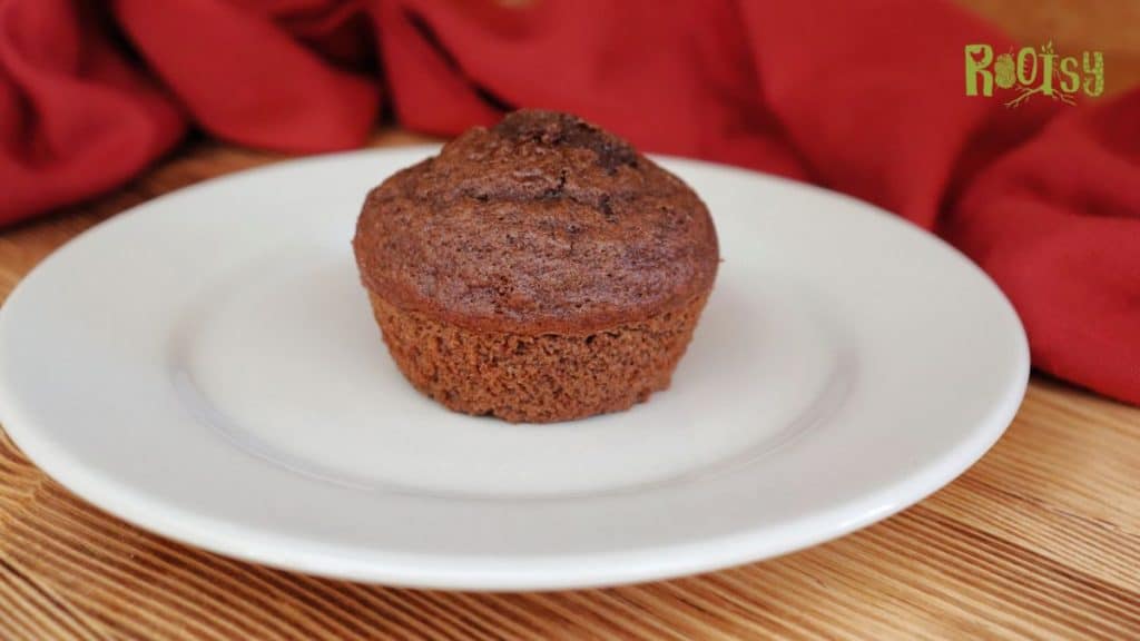 A single chocolate muffin sits on a white plate with a wooden surface and a red cloth in the background. The word "Rootsy" is visible in the top right corner.