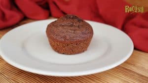 A single chocolate muffin sits on a white plate with a wooden surface and a red cloth in the background. The word "Rootsy" is visible in the top right corner.