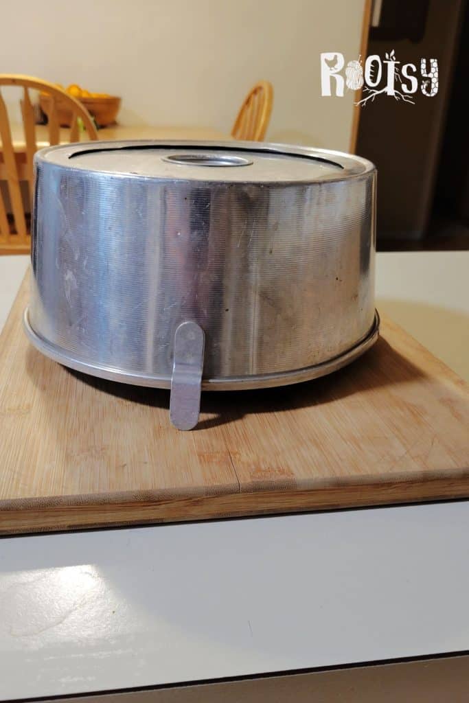 An upside-down metal angel food cake pan is resting on a wooden cutting board on a kitchen counter. A wooden chair and a bowl of fruit are visible in the background. The word "Rootsy" is in the top right corner.