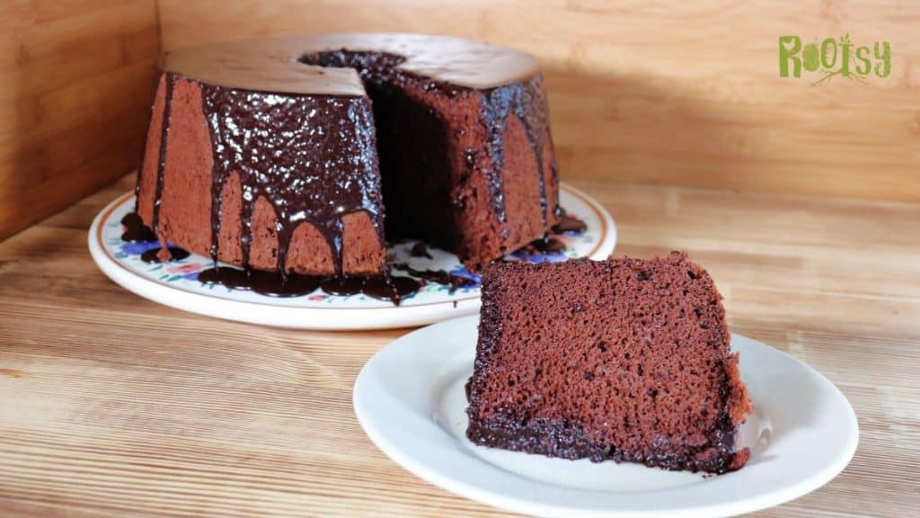 A rich chocolate bundt cake with glossy chocolate glaze sits on a plate, with a large slice cut and placed on a smaller plate in the foreground. The background features wooden surfaces and a “Rootsy” logo.