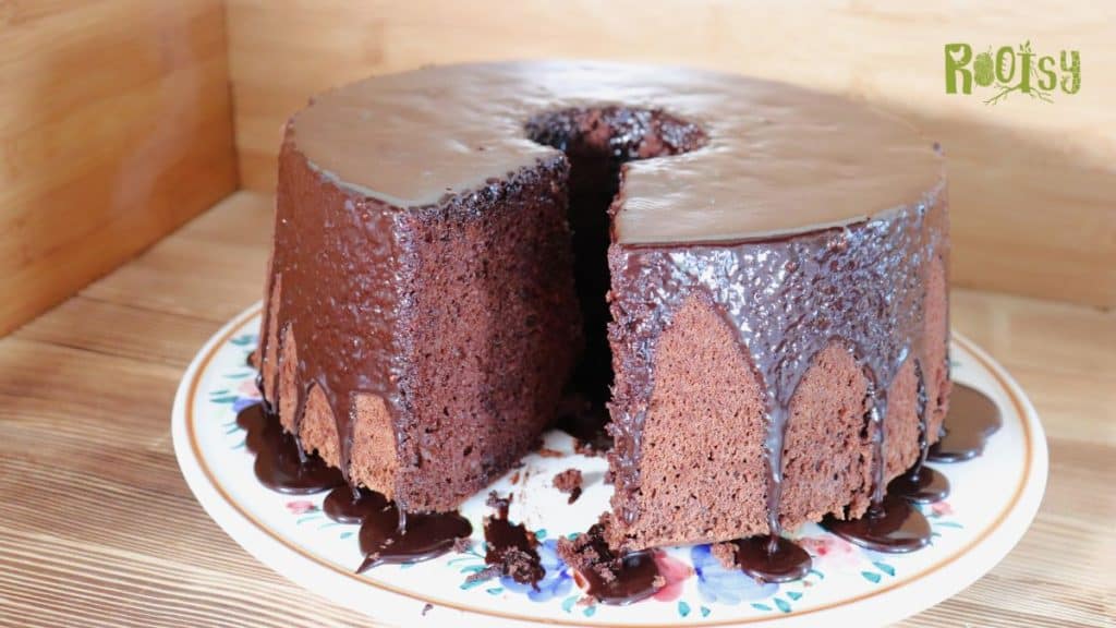 A tall, round chocolate cake with a hole in the center sits on a plate. One large slice has been removed, revealing the moist interior. Chocolate glaze drips down the sides and onto the plate.