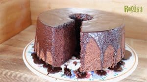 A tall, round chocolate cake with a hole in the center sits on a plate. One large slice has been removed, revealing the moist interior. Chocolate glaze drips down the sides and onto the plate.