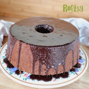 A round chocolate chiffon cake with a hole in the center sits on a floral plate, topped with glossy chocolate glaze dripping down the sides. The background is wooden, and “Rootsy” is written in green at the top right.