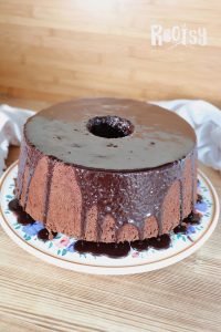 Chocolate chiffon cake with a shiny chocolate glaze dripping down the sides, displayed on a floral-patterned plate against a wooden background.