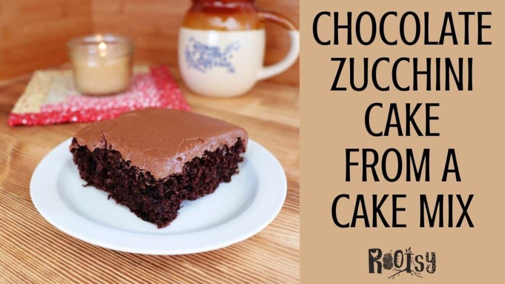A slice of chocolate zucchini cake with frosting sits on a white plate. In the background, there’s a candle, mug, and red cloth. Text reads: "Chocolate Zucchini Cake from a Cake Mix - Rootsy.
