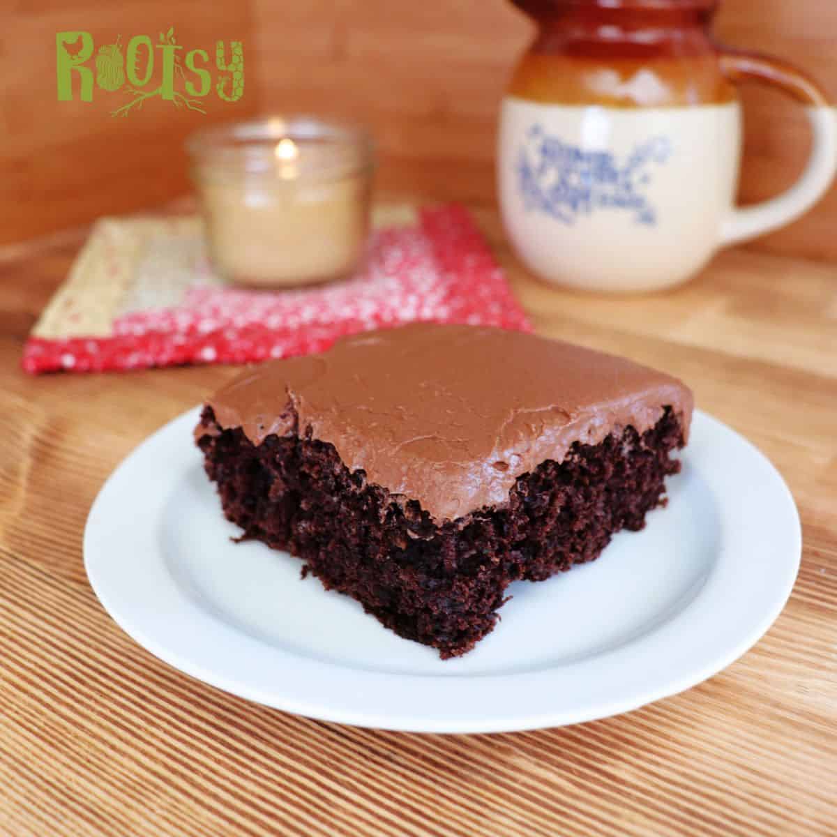 A slice of chocolate cake with thick chocolate frosting sits on a white plate. In the background, there is a mug, a candle, and a red patterned napkin on a wooden surface. The word "Rootsy" is in the top left corner.