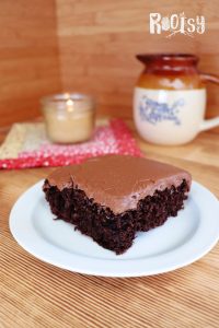 A slice of chocolate cake with chocolate frosting sits on a white plate. In the background, there is a candle, a patterned mat, and a ceramic mug on a wooden surface. The word "Rootsy" appears in the top right corner.