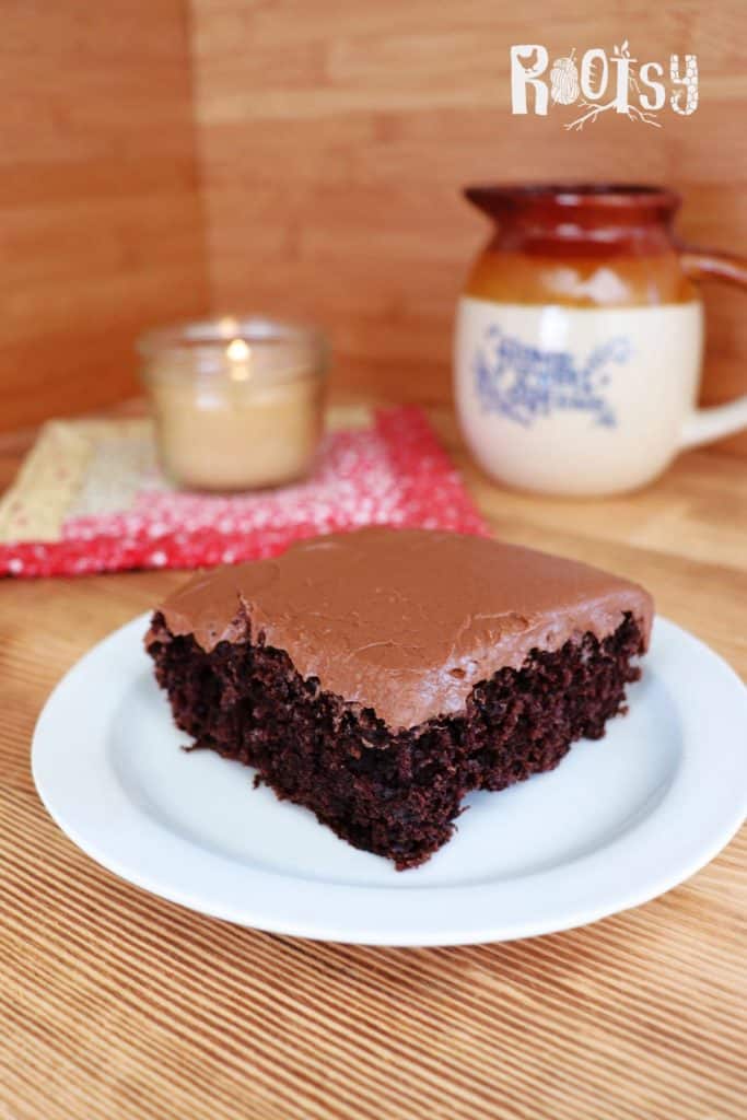A slice of chocolate cake with chocolate frosting sits on a white plate. In the background, there is a candle, a patterned mat, and a ceramic mug on a wooden surface. The word "Rootsy" appears in the top right corner.